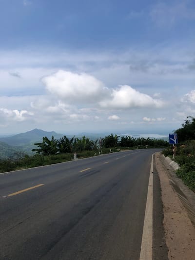 The image captures a winding asphalt road on a mountainside overlooking a valley and distant body of water, possibly a reservoir, near Phan Son, Vietnam. The road is marked with a yellow dashed line and a white solid line on the right edge. Lush green vegetation, including banana trees, lines the roadside, with some scattered rocks and dirt. In the distance, rolling hills and mountains are visible under a partly cloudy blue sky. A blue traffic sign with Vietnamese text and directional arrows is posted on the right side of the road, indicating a sharp bend. There are no people or animals visible in the scene.