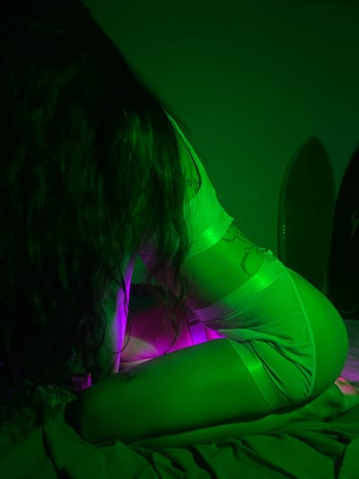 The image depicts a woman in a sexually suggestive pose in an interior space. She is bent over, with her back arched and buttocks prominent. She is wearing white underwear. Her long dark hair obscures much of her face. The lighting is green and purple. In the background, there is a blurred object resembling a ceiling fixture. The image was supposedly taken in Heenatigala Junction, Sri Lanka.