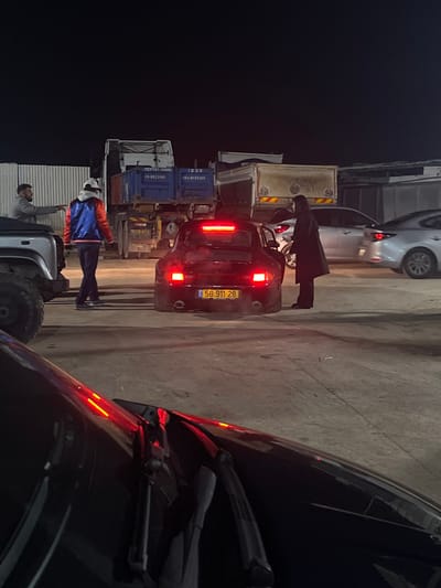 This nighttime image, captured in Kfar Qasim, Israel, depicts a scene in what appears to be an industrial or commercial loading area. The photograph is taken from inside a dark vehicle, with its hood and windshield wipers partially visible in the right foreground.

In the mid-ground, a black car with its brake lights illuminated is prominent. Its yellow Israeli license plate clearly displays the number **58-911-28**. A person dressed in a long dark coat or abaya stands near the rear of this black car, possibly attending to the trunk or simply standing beside it. Behind this black car, a silver or light-colored sedan is partially visible.

To the left of the black car, a quad bike or ATV is parked. Two other individuals are seen near it: one in a vibrant blue and orange/red jacket, and another with a beard wearing a grey top. They appear to be casually standing or interacting.

The background features several large trucks, possibly dump trucks or flatbeds, with blue containers or beds. Some visible text on these containers includes a phone number "053-851-..." and possibly "GENERAL CARGO" on one, and "050-8020101" on another. The ground is concrete, typical of such industrial settings.

The overall atmosphere suggests a quiet moment at night in a functional, non-residential area, with vehicles and people present, possibly for work-related activities or a stopover.