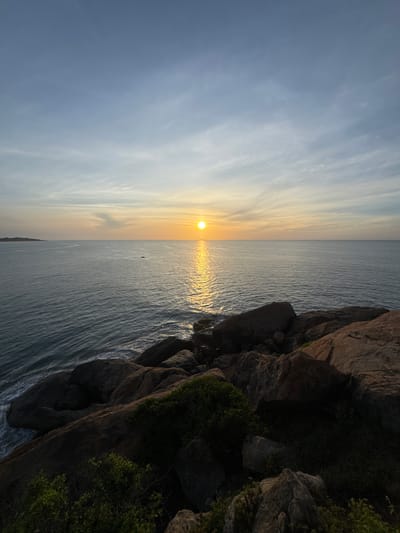 The image captures a serene coastal scene at sunset, with the sun low on the horizon, casting a golden glow across the calm sea. The sky is a soft gradient of blue and yellow, with wispy clouds suggesting fair weather. To the right of the frame, rugged, dark brown rocks rise up, partially covered by lush green foliage, indicating a rocky coastline. The sea itself is a deep blue-gray, with gentle ripples reflecting the sunlight, creating a shimmering pathway towards the horizon. A solitary, small dark object, possibly a boat or a person on a board, is visible in the distance on the water. The overall mood is peaceful and tranquil, evoking the beauty of a sunset over the ocean. The image is oriented vertically, suggesting it was taken from a high vantage point looking down towards the sea and sky. No text is visible in the image.