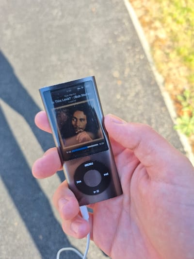 The image shows a person's hand holding an Apple iPod with the song "Is This Love?" by Bob Marley playing. The iPod is black and silver, and the screen displays the song title, artist, and album cover. The song is currently at 0:42. The person is standing on a sidewalk with a patch of grass to the right. The coordinates provided correspond to a location in East Hertfordshire, United Kingdom. The specific location is a residential area with homes and streets.
