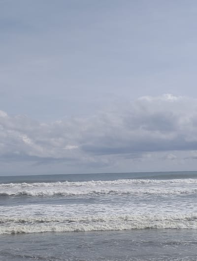 The image depicts a serene beach scene in Meudheun, Indonesia. Gentle waves with white foam are rolling onto the shore under a cloudy sky. The horizon is a soft blend of the ocean and the sky, with diffused sunlight filtering through the overcast conditions. No people or man-made objects are visible, emphasizing the natural beauty of the coastal environment. The water appears calm, and the overall atmosphere is tranquil and expansive.