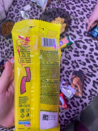 The image displays a close-up of a yellow package of Chupa Chups candy, held by a person's hand. The package features prominent branding and nutritional information. The background is blurred, but suggests a domestic setting with a leopard-print fabric and other objects. There is no visible indication of the city of Zmeinyi, Russia, in the image. The lighting appears to be natural, possibly from a window.