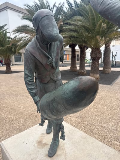 A bronze statue stands on a concrete pedestal, set against a background of palm trees and urban architecture. The statue depicts a figure with a head covering, possibly a turban, wearing a buttoned jacket. The figure is mounted on a large, indistinct object, potentially a creature or vehicle. The location is likely Arrecife, Spain, suggested by the type of architecture and foliage. The environment includes a tiled pavement and buildings in the background.