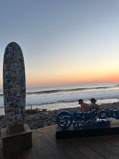 A sunset view of El Salvador’s coastline, featuring gentle waves. Two people stand near a blue sign. A surfboard covered in stickers rests on a base in the foreground. The sky blends from blue to orange near the horizon. A wooden deck and rocks add texture to the scene.