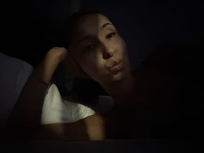 A dimly lit, close-up, horizontal, medium shot photograph shows a woman's face, with her head resting on her arm, as she looks at the camera. Her face is partially obscured by shadow, but a sliver of light illuminates her right eye, cheekbone, and lips. The woman has dark hair pulled back and is wearing light-colored foundation, dark eyeliner, and mascara. Her lips are slightly parted, and she appears to be pouting. The background is dark and blurry, with a hint of white fabric, possibly a pillow or sheet, on the left side of the frame. The overall mood of the image is intimate and slightly mysterious due to the low lighting. The location of Doma 1096 Km, Russia is not visible in the image.