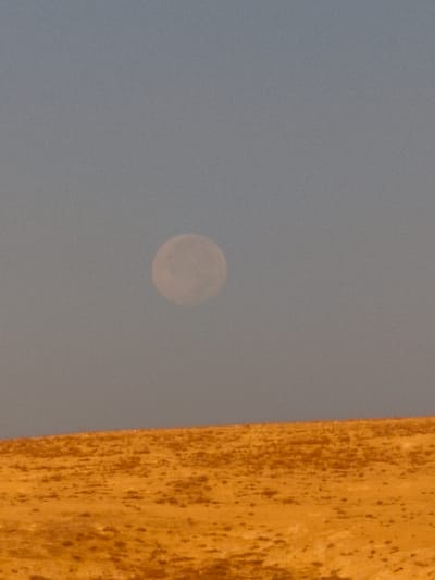 A full moon is visible in the hazy, light blue sky above a sandy, orange-hued desert landscape. The moon is positioned slightly to the left of the center of the image. The desert terrain in the foreground is uneven, with a gentle slope rising towards the sky. Small, dark specks dot the surface of the desert, possibly indicating sparse vegetation or rocks. The overall impression is one of a vast, arid environment under a diffused sky.