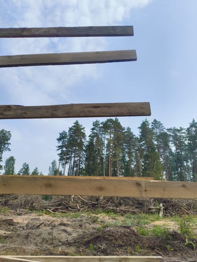 In Дашкаўскі сельскі Савет, Belarus, an outdoor scene depicts the construction of a wooden structure. The foreground features several horizontally placed wooden beams, some of which are unfinished and weathered. In the background, a dense forest of tall pine trees stretches across the horizon, with a patch of sky visible above. The ground is a mixture of sandy soil, dirt, and scattered debris, suggesting a construction or clearing site. The overall impression is one of an unfinished building project amidst a natural woodland environment.