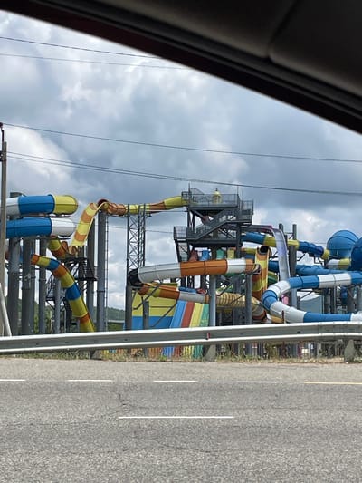 The image shows a complex structure of water slides at an amusement park in Lermontovo, Russia. The slides are made of brightly colored plastic tubes, predominantly in yellow, orange, blue, and white. They twist and turn at various heights, supported by a metal framework. Some slides are open tubes, while others appear to be enclosed. There are visible staircases and platforms associated with the slides, indicating multiple levels and entry points. The overall impression is one of a large, elaborate water park. The sky above is cloudy and overcast, suggesting a potentially humid or rainy day. In the foreground, a road with white lane markings is visible, separated from the park by a metal barrier. The environment appears to be a mix of park infrastructure and natural elements, with some greenery visible behind the slides. There are no people clearly visible in the image.