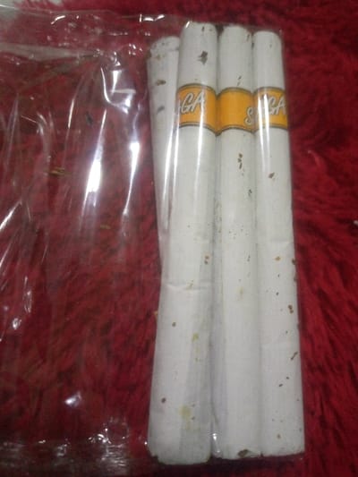 A close-up, top-down view captured in Kuningan, Indonesia, displays four white, cylindrical objects resembling large kretek cigarettes or cigars, packaged together within a clear, somewhat wrinkled plastic wrapper. Each object is adorned with a prominent yellow band bearing the word "SAGA" in white or light-colored text. The white surfaces of the cylindrical items exhibit numerous small, irregular brown and yellowish flecks. The packaged items rest on a textured, deep red surface, likely a fabric or carpet. No individuals are present, and no actions or interactions are depicted within the static scene.