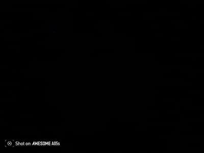The image is predominantly black, indicating a very dark or underexposed scene. There is a small, faint blue speck visible in the upper portion of the image. In the bottom left corner, a white text overlay reads "Shot on AWESOME A05s" with a circular icon to its left. The scene appears to be indoors or at night due to the lack of ambient light. There are no discernible people, subjects, objects, actions, or environmental details beyond the text overlay and the single blue speck. The location in Alalubosa, Nigeria is not depicted in the visual information provided by the image.