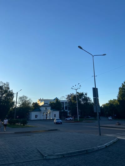 The image depicts a street scene in Nyzhni Mlyny, Ukraine, on a clear day. The sun is low in the sky, indicating it is either early morning or late afternoon, casting long shadows. The scene is divided by a clear blue sky on the left and a street with a paved sidewalk on the right.

Several streetlights are visible, with a large double-headed one dominating the left side of the frame. Traffic lights and road signs are mounted on a pole near the center. In the background, there are buildings with white facades and green roofs, surrounded by lush green trees. A few cars are on the road, including a blue car and a white one.

On the paved sidewalk, two people are walking, one in a white t-shirt and black shorts, the other in dark clothing. They appear to be engaged in a casual walk. The overall atmosphere is peaceful and tranquil, suggesting a normal day in the town. There is no visible text in the image.