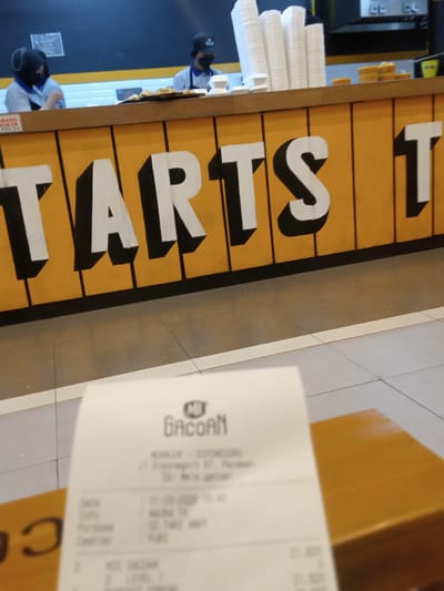 The image depicts the counter of a Mie Gacoan restaurant in Nganjuk, Indonesia. The counter's facade is painted with the word "STARTS" in large, bold letters. Behind the counter, two individuals wearing face masks and head coverings are visible, possibly employees. Stacks of white disposable cups and possibly food items are arranged on the counter. In the foreground, a receipt from Mie Gacoan is placed on a wooden surface. The receipt contains information such as the date (21-03-2023), branch location (Nganjuk - Diponegoro), and order details.