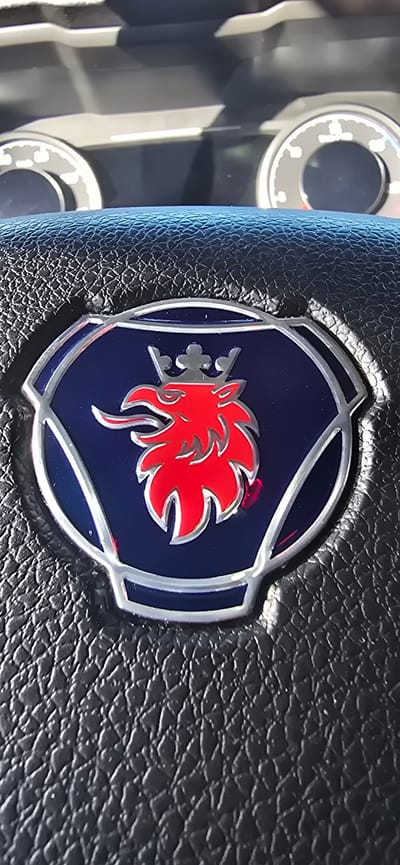 This is a close-up, vertical, eye-level shot of the steering wheel of a Scania truck. The central focus is the Scania logo, a red griffin with a crown on a dark blue background, encased in a silver emblem. The steering wheel itself is made of black, textured leather. In the upper portion of the image, out of focus, parts of the truck's dashboard are visible, including two circular gauges. The lighting suggests it is daytime, possibly with sunlight reflecting off the gauges. The image was taken inside a Scania truck, location unspecified beyond the city of Pedrola, Spain.