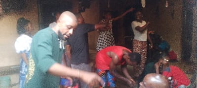 A group of people is gathered indoors, possibly for an event. A man in a green shirt is in the foreground. Behind him are several other individuals in casual clothing. One man is wearing a red tank top and colorful shorts, bending over. Two women in the background have their arms raised. The setting appears to be a modest home or community space, with simple architecture. The lighting is dim.