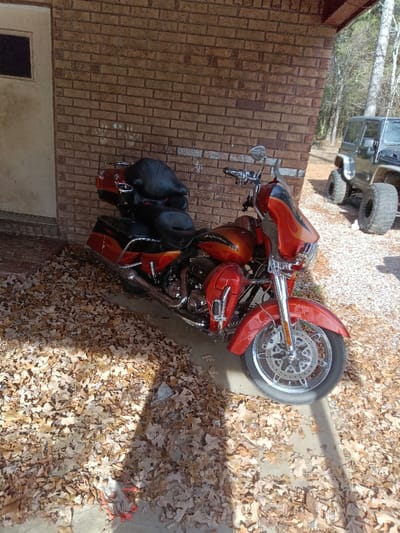 An orange and black motorcycle is parked on a concrete pad covered in brown leaves. The motorcycle is next to a brick wall, and a gray Jeep with large tires is visible in the background. The scene appears to be outdoors, possibly in a yard or driveway during the autumn season. The overall lighting suggests a sunny day.