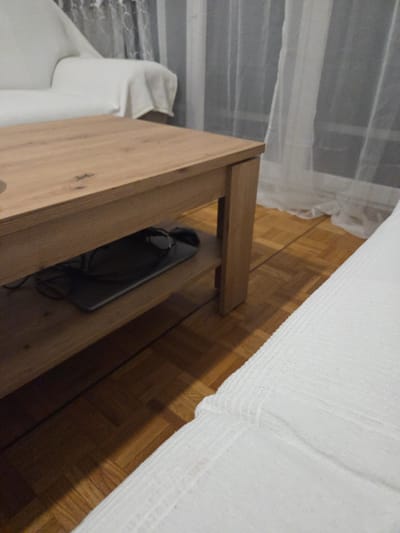 This image depicts an indoor residential scene, likely a living room, located in Lausanne, Switzerland. No individuals are present, and no actions or interactions are occurring.

In the immediate foreground, a white, textured fabric, consistent with a sofa or bed cover, occupies the lower right quadrant of the frame. In the midground, a rectangular light wood coffee table with a lower shelf is centrally positioned. On this lower shelf, a dark-colored tablet or similar electronic device lies flat, with dark cables or headphones resting on its surface.

To the left of the coffee table, a white upholstered sofa is visible, with a white throw draped over its armrest. Behind the coffee table and to the right, a window is partially visible, covered by translucent white sheer curtains. The floor throughout the visible area is covered with light brown parquet flooring arranged in a herringbone or similar geometric pattern. Ambient lighting creates shadows on the floor.