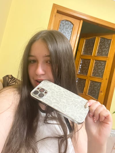 A woman with long brown hair is holding a smartphone up to her face, partially obscuring her mouth. The phone has a white patterned case with three visible camera lenses. She has painted fingernails with small red hearts on them. The background includes a yellow wall and a wooden-framed door with decorative glass panels. There is no information available indicating the location is Cherliany, Ukraine.