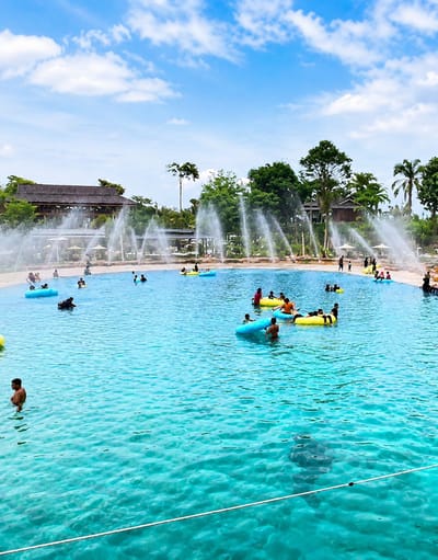 The image shows a crowded swimming pool at a water park in Eampat Kwalamencirim, Indonesia. Numerous people are visible in the clear turquoise water, many of whom are floating on inflatable tubes of various colors, including yellow and blue. Some individuals are swimming, while others are sitting or lying on the inflatables. In the background, a large building with a traditional wooden roof structure is situated behind a line of trees. Several fountains are spraying water upwards, creating a mist effect in the air. The sky above is blue with scattered white clouds. The overall scene depicts a lively and active recreational environment.