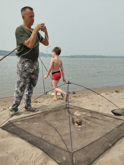 An adult male wearing a green t-shirt and camouflage pants is standing on a sandy beach near a body of water. He is holding a fishing rod and appears to be untangling some fishing line. A young boy, wearing red swim shorts, is walking away from the man towards the water. In the foreground, a fishing net lies open on the sand, with a small object, possibly bait, inside. Fishing rods are visible in the background, along with a distant treeline and a hazy sky. The scene is consistent with a fishing activity on the shore of the Ob River in or near the village of Bibikha, Russia.