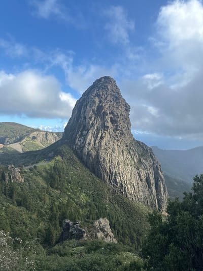 A prominent, colossal rock formation, identified as Roque de Agando, dominates the central and right portions of the frame within the mountainous landscape of La Gomera, Spain, in the vicinity of San Sebastián de la Gomera. The rock is characterized by its steep, rugged, vertically striated faces, ranging from light gray to tan, and culminates in a sharp peak. Lush, green vegetation, consisting of trees and shrubs, covers the lower slopes and surrounding terrain, extending into the foreground. A dark, winding paved road is visible on a vegetated ridge to the left of the primary rock formation. Distant, hazy mountain ridges recede into the background. The sky is predominantly blue with scattered white cumulus clouds. No human figures or active interactions are present.