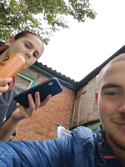 A low-angle, close-up selfie shows a young woman holding an orange can and a dark green smartphone in her left hand. She is wearing a dark t-shirt. To her right, a young man's face is visible in the foreground, wearing a blue jacket. Behind them, a brick building with a green window frame is visible, along with a wooden structure and corrugated metal roofing. Trees with green leaves are overhead. The background appears to be an outdoor setting, possibly a yard or a rural area, consistent with locations in Okhtyrka, Ukraine.