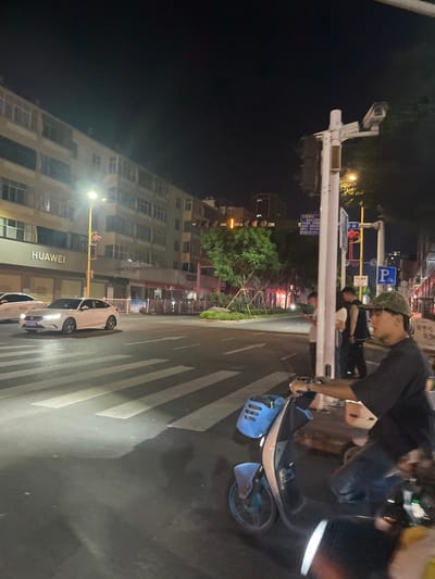 The image depicts a nighttime street scene at an intersection in Dali City, China.

In the immediate foreground, to the right, a person is seated on a light blue electric scooter. This individual, appearing male, wears a dark t-shirt, dark pants, and a camouflage-patterned cap. Their right hand is on the scooter's handlebar. The scooter features a blue storage basket on its front. A pedestrian crosswalk, marked by white stripes, extends across the asphalt road in front of the scooter.

In the midground, a white four-door sedan is positioned on the road, facing left, with its headlights illuminating the crosswalk. A second white vehicle is partially visible behind it. To the left, a multi-story building houses a "HUAWEI" retail store on its ground floor, marked by a prominent white sign. A street light with a red traffic signal is visible near this building.

At the center of the intersection, a small landscaped island contains green trees and shrubbery. Multiple traffic light poles and street signs, displaying Chinese characters, are visible throughout the scene. One tall, white traffic light pole on the right side of the image features a mounted camera. Another traffic signal further down the street displays a yellow or amber light.

On the sidewalk to the right, near the traffic light pole, two individuals are standing. One person, wearing a light-colored shirt, appears to be looking at a mobile phone. The background consists of additional multi-story urban buildings under the dark night sky. Artificial lighting from streetlights and building facades illuminates the environment.