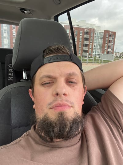 A man wearing a brown t-shirt and a black baseball cap is in the driver's seat of a car. He has a beard and is resting his arm on the headrest. In the background, a brick building with multiple windows and balconies is visible. This building is located in the port area of Wilhelmshaven, Germany, specifically near the new building complex called "Speicherinsel".