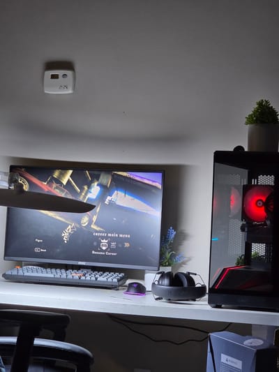 This media file shows a desk setup with a computer, monitor, keyboard, and headphones. The computer tower has a red light on the fan, and the monitor displays a video game menu titled "career main menu" with the option to "Resume Career". A knife is placed in front of the monitor, its blade pointing upwards. A small plant is visible to the left of the computer, and a lavender plant is behind the headphones. The desk is illuminated by artificial light. The background appears to be a wall. A warning label is visible on a box to the right of the computer.