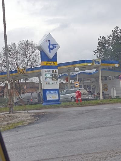The image depicts a Petrol branded gas station located in Radomirtsi, Bulgaria, under an overcast sky. A prominent blue and white price totem displays "perfekt" branding, the Petrol logo (a stylized bird), and fuel prices: DIESEL 1.61, 95 1.4, and BLUE 0.6. The totem also features Bulgarian text "ЗАРЕДЕТЕ ПРИ НАС" (Fill up with us).
The gas station features a blue and yellow striped canopy covering fuel pumps, beneath which a silver station wagon is parked. A darker grey panel van with its rear passenger-side door open is parked on a grassy area to the left of the main entrance. A white vehicle is partially visible behind the silver station wagon. Two individuals are seated on a bench or similar structure near the gas station building, to the right of the silver station wagon.
Additional signs visible include a "DKV" sign under the canopy and a red hexagonal "СТОП" (STOP) sign on a pole near the silver station wagon. The scene is viewed from a paved road in the foreground. A utility pole stands on the left. In the background, trees, some bare and some evergreen, along with residential structures, are visible under the grey sky.