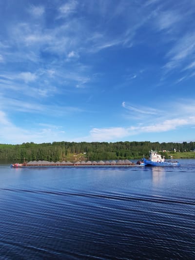 A wide shot captures a blue and white barge, pushed by a blue tugboat, transporting a long line of gray cargo containers along a body of water. The water is deep blue and rippled with waves. The sky is bright blue with wispy white clouds scattered across the expanse. Along the shore of the body of water, a green tree line runs horizontally across the middle of the image. On the right side of the tree line, there is a small cluster of houses. The location may be in the city of Ankimarovo, Russia.
