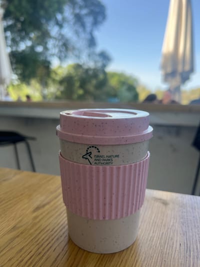 A reusable travel mug, light beige with pink speckles, a matching pink lid, and a ribbed pink silicone sleeve, rests on a light wooden table with visible grain. The mug features a black logo for the 'ISRAEL NATURE AND PARKS AUTHORITY,' depicting an ibex head. In the soft-focus background, green trees and a clear blue sky are visible, alongside a closed light-colored umbrella on the right and blurry figures of people. A dark chair is partially visible on the left. The scene is set outdoors, likely at a cafe or visitor center associated with a national park in Rosh Haayin, Israel.