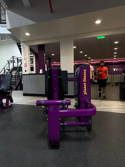 The image depicts the interior of a "Planet Fitness" gym located in Nuevo Santiago, Panama. In the foreground, a large, purple and black resistance exercise machine, likely a leg press or similar, is centrally positioned on a dark, speckled rubberized gym floor. Its side panel displays "planet fitness" vertically in yellow text. To the far left, another purple and black exercise machine is partially visible.
The midground features two adult males walking from right to left. The man in the foreground is wearing a red sleeveless shirt, dark shorts, and athletic shoes, holding a black water bottle in his right hand. Behind him, another man wears a light pink short-sleeved shirt. The flooring transitions from the dark rubberized surface to a lighter, wood-look laminate or tile in the background.
The gym's interior design includes white structural columns with faint grey gear patterns, purple accent walls, and a high ceiling with recessed circular lighting and linear fixtures. A green exit sign is visible on the ceiling in the upper right. To the far left, a black metal staircase ascends to an upper level, partially obscuring white walls with black text, partially legible as "you be". A red fire extinguisher is mounted on a column near a grey counter. In the far background, a "TRX" sign is visible on a wall, and a black mesh trash can is partially seen on the right.