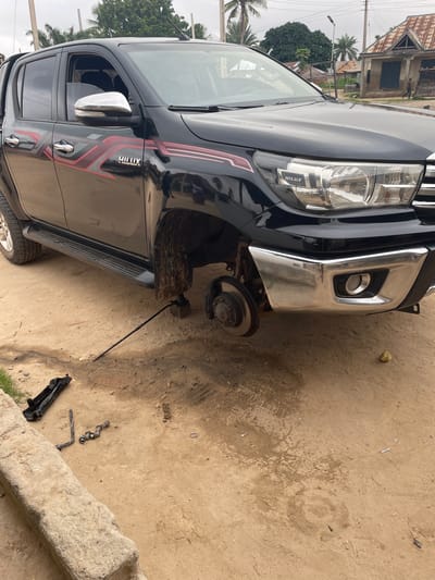 The image depicts a black Toyota Hilux pickup truck with its front passenger-side wheel removed, being supported by a jack. The removed wheel and lug nuts are not visible. A black jack handle and a set of lug nuts are scattered on the sandy ground near the truck. The truck is parked on a dirt road in Kwalla, Nigeria, indicated by the presence of a residential building with a corrugated metal roof and the general environment. The sky is overcast.