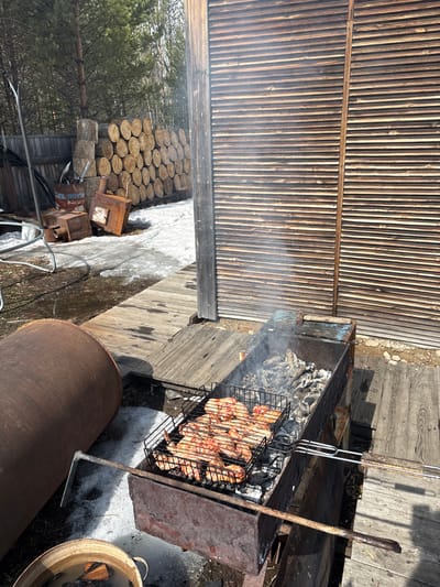 The image depicts a barbecue scene in what appears to be a backyard or outdoor area, possibly located in a rural or suburban setting.
**Objects:**
* A barbecue grill with meat being cooked on a wire rack over hot coals.
* Grilling tools
* Wood logs stacked to the side.
* A metal pipe or cylindrical object.
* Wooden planks making up a deck or platform.
* A shed-like structure with wooden slat walls.
* A bucket containing water and wooden scraps
**Environment:**
* The presence of snow suggests a winter or early spring season.
* Trees visible in the background indicate a location near a wooded area.
Given the request to include the location in Zheleznodorozhnyi, Russia, it is possible that this scene is occurring in the area surrounding this city, possibly in a dacha or private property outside the urban center.