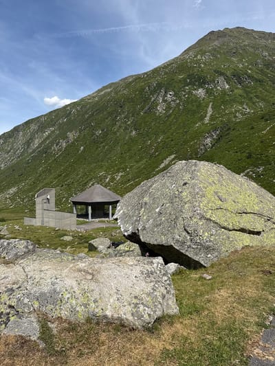 In Hospental, Switzerland, a mountain landscape is depicted under a partly cloudy sky. In the foreground, large, lichen-covered boulders rest on a grassy slope. Behind them, a modern concrete structure stands to the left, featuring a vertical tower with antennas and a lower, angled section. To the right of the concrete building, an open-sided pavilion with a thatched roof is situated on a paved area. The background is dominated by a steep, green mountain slope dotted with rocks and vegetation. A thin contrail is visible in the sky.