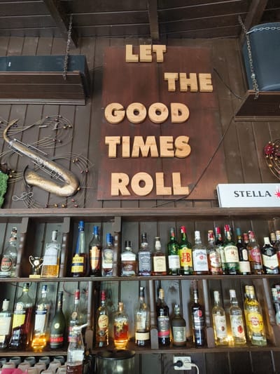 The image shows a wall in a bar or restaurant in Târgoviște, Romania, decorated with wooden letters spelling "LET THE GOOD TIMES ROLL." Below the sign, shelves are stocked with various bottles of alcohol. To the left of the sign, a metal sculpture resembling a saxophone and musical notes is mounted on the wall. The overall ambiance suggests a casual and possibly vintage-themed establishment. The lighting is warm and appears to be artificial, highlighting the bottles on the shelves. No people are visible in the image.