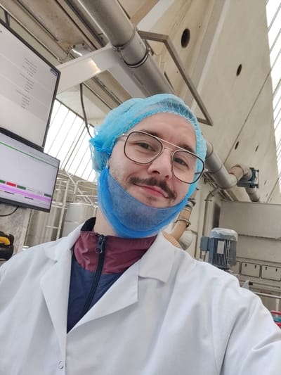 A selfie taken by a Caucasian male with a mustache, wearing glasses, a blue surgical cap and mask, and a white lab coat over a maroon and blue jacket. He is in an industrial environment, possibly a factory, with exposed pipes, machinery, and a control panel with a screen displaying data. The background features large windows that let in natural light. The city of Oosterhout, Netherlands, cannot be confirmed as the location based on the image.