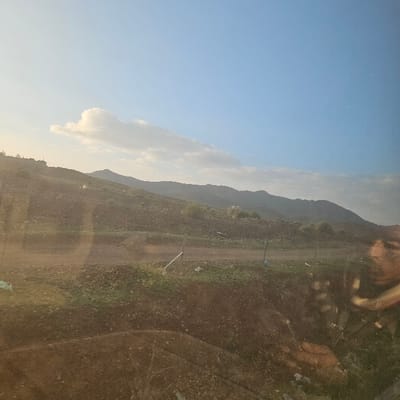 The image depicts a rural, mountainous landscape in Ouahat Sidi Brahim, Morocco, viewed through a reflective surface, likely a vehicle window. The sky is a clear, light blue with an elongated white cloud formation in the upper left quadrant and fainter clouds along the distant horizon. The terrain consists of dry, rocky, undulating hills and mountains receding into the background.
In the mid-ground, a horizontal dirt path or unpaved road traverses the scene. Sparse patches of green vegetation, consistent with scrub or small plants, are interspersed among the brown earth and scattered small rocks. Several thin, light-colored poles are vertically embedded in the ground adjacent to the path. A faint, light-colored structure is visible on the crest of a hill on the far left.
The foreground is dominated by darker, disturbed earth, suggesting a roadside embankment or ditch. Prominent reflections are superimposed over the landscape, most notably a distorted reflection of a human face and upper torso on the right side of the image. No individuals are physically present within the landscape itself.