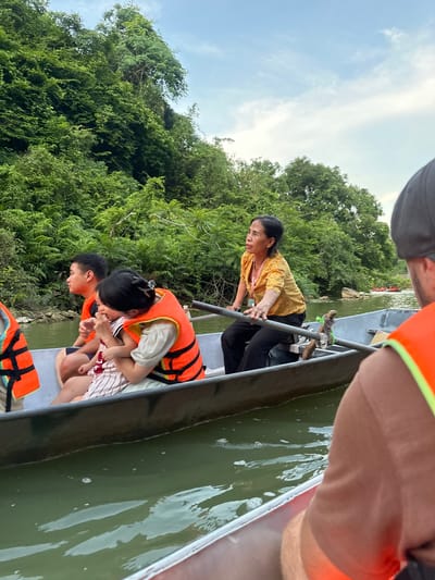 In the foreground, the back of a person wearing an orange life vest is visible. To their left, a woman with a yellow patterned shirt and black pants is rowing a boat with a wooden oar. In the boat with her are a young boy wearing an orange life vest and a woman holding a baby. The boat is on a body of water, with a lush green, tree-covered hillside on the left. The sky above is a pale blue with scattered clouds. Further in the distance, other boats can be seen on the water. The scene appears to be a scenic river or lake in Yên Sơn Ward, Vietnam, likely a tourist or recreational activity.