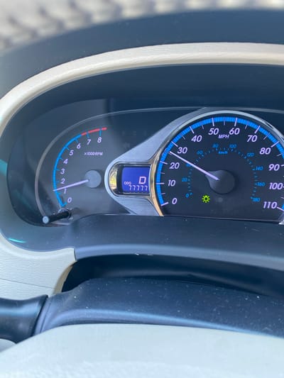 This is a close-up, eye-level, photograph of a car's dashboard, specifically the speedometer and tachometer. The dashboard is predominantly black with some light beige trim. The speedometer, on the right, is marked in MPH and KM/H, with the needle indicating a speed between 30 and 40 MPH, closer to 30 MPH. It also shows a green indicator light, possibly for the headlights or daytime running lights. The tachometer, on the left, is marked in "x1000 RPM" with the needle indicating a speed between 0 and 1, suggesting the engine is idling or at very low RPM. In the center-right of the image, a digital display shows "ODO" followed by the number "77777" which is likely the odometer reading. The image does not contain any information about the city of Prijeradi, Montenegro.