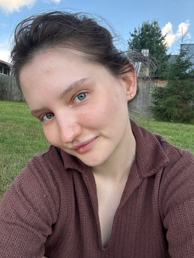 A young woman with light skin, light blue eyes, and dark brown hair is pictured outdoors in a grassy area. She is wearing a loose-fitting, maroon-colored knit top. Her hair is pulled up and appears windswept. She has a small stud earring in her left ear. In the background, a wooden fence, trees, and a cloudy blue sky are visible. The location is described as being in the city of Burdyata, Russia. The lighting suggests it is daytime.