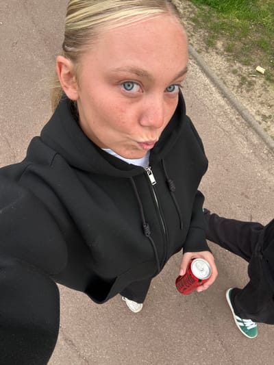 A woman takes a selfie while walking down a street with a person. The woman has blonde hair, fair skin, and blue eyes. She is wearing a black hoodie with a white t-shirt underneath. She holds a red can with a silver top in her right hand. In the background, a person walks with the woman. This person wears black pants and green and white Adidas sneakers. The sidewalk is made of gray concrete and has some green grass. The location is unknown.