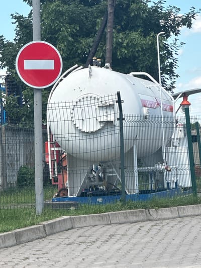 The image shows a large, white, cylindrical storage tank, likely a tanker truck or silo, located behind a wire fence. The tank has some red and white lettering on its side, but it is difficult to read. There is also an orange conical object at the top of the tank. In the foreground, a circular red and white traffic sign with a white rectangle in the center is visible, indicating "Do Not Enter." The scene appears to be outdoors, with greenery and trees in the background and a paved area to the right. The sky is blue with some clouds, suggesting daytime weather. The overall impression is of an industrial or logistical area, possibly a storage facility or transport hub. There is a hint of a person's face peeking out from behind the tank in the lower right portion of the image, obscured by the fence.