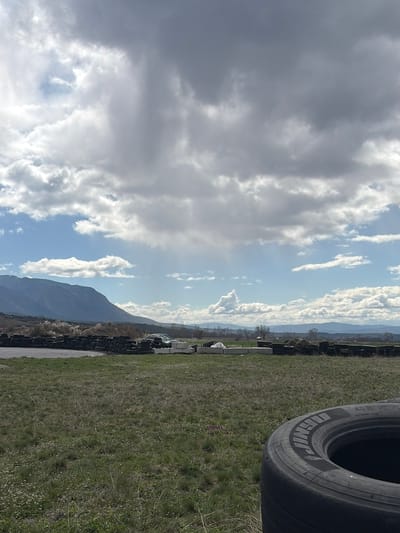The image shows an outdoor scene near Kraynitsi, Bulgaria. The foreground is dominated by green grass. A stack of tires is present on the right. In the background, there is a mountain range beneath a sky with varied cloud cover.