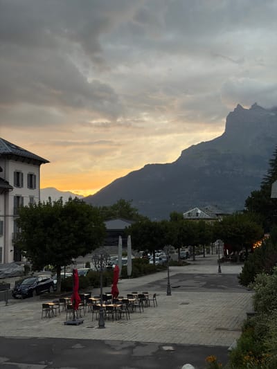 The image captures a view in Saint-Gervais-les-Bains, France, featuring a street scene at dusk with a mountain range in the background. On the left, a multi-story building with a light-colored facade, white trim, and gray shutters is visible. Adjacent to the building are trees. In the foreground, an outdoor seating area with tables and chairs is set up, some covered by red umbrellas. The pavement is a patterned cobblestone. A dark-colored car is parked to the left of the seating area. The street continues into the distance, lined with trees and further buildings, and is partially visible with cars parked along it. The sky is filled with dramatic clouds, with the setting sun casting a warm glow over the horizon and silhouetting the mountains. The overall impression is of a quiet evening in a mountain town.
