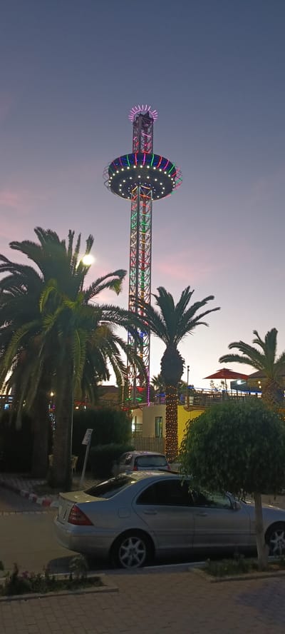 The image depicts an amusement park attraction, possibly a drop tower, at dusk in Hammam Sousse, Tunisia. The tower is illuminated with colorful lights, and a circular platform at its top is also lit. Palm trees are visible in the foreground, with one of them decorated with string lights. Two cars are parked on the street in front of the amusement park. The sky is a gradient of purple and blue, indicating the transition from daylight to night. The scene suggests a lively atmosphere at an entertainment venue.