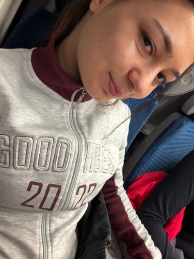 A young woman with dark hair and eyes poses for a close-up selfie while sitting in an airplane seat. She wears a light gray sweatshirt with a burgundy collar and trim. The sweatshirt features the words "GOOD VIBES 20" in a blocky, maroon font. The seat behind her is upholstered in blue fabric. Another person, wearing black pants, sits next to her, with a red garment visible between them.