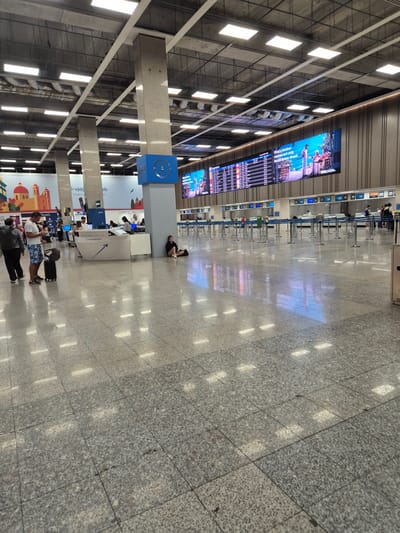 The image depicts the interior of an airport terminal, likely the passenger departure area, in Luqa, Malta. The scene is characterized by a vast, polished granite floor reflecting the overhead lighting and large digital display screens. Several people are visible, some standing and interacting at a customer service desk, while others are seated or walking with luggage. A woman is seen sitting on the floor, leaning against a pillar, near the information desk. The departure board displays flight information, and queues of people are absent from the check-in counters, suggesting a less busy period or an area not yet in full operation. The architectural style includes large concrete pillars supporting a high ceiling with numerous fluorescent lights. The overall impression is one of modern airport infrastructure.