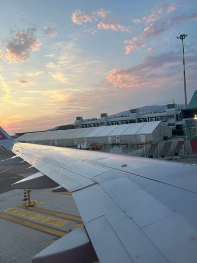 This media file depicts an airplane wing in the foreground, casting a shadow on the tarmac at Lamezia Terme, Italy. In the background, airport infrastructure is visible, including a terminal building with numerous air conditioning units, and a large tent-like structure. Beyond the airport, mountains are faintly visible under a twilight sky. The sky itself is a dramatic spectacle of sunset colors, with hues of pink, orange, and yellow blending into the blue. Wispy clouds are scattered across the sky, catching the light of the setting sun. The overall mood is serene and anticipatory, suggesting the calm before a flight or the end of a day of travel. Visible text includes "E175" and what appears to be a flight number or gate information on the tarmac markings. The scene captures a moment at an airport during a beautiful sunset.